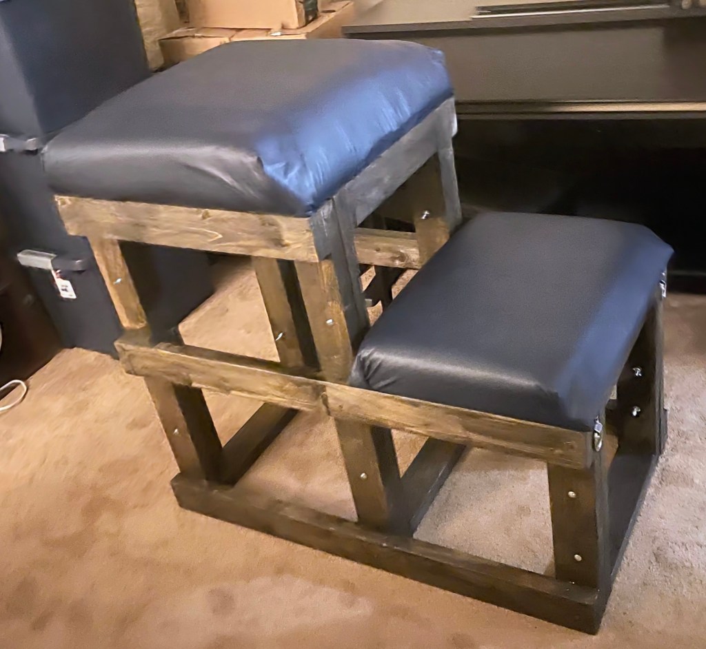 A spanking bench, shaped like a set of two stairs, with a wooden frame and squishy blue cushions for the upper body and knees.