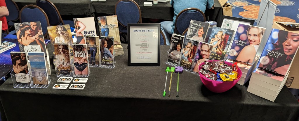 My sales table at Frolicon 2023. From left: Shell Game, Lessons, Butt Stuff, Baker's Dozen, Training the Trainer, Dreaming of How it Was Going to Be, Weekend Plans, and Switching Plans. In the front row: flyers for all of those books plus Other Options and Three Paddlings for Kristen, AASS (Atlanta Area Spanking Society) cards, a dick stick, a Lego evil stick, and a unicorn evil stick, and then a bowl of M&Ms.