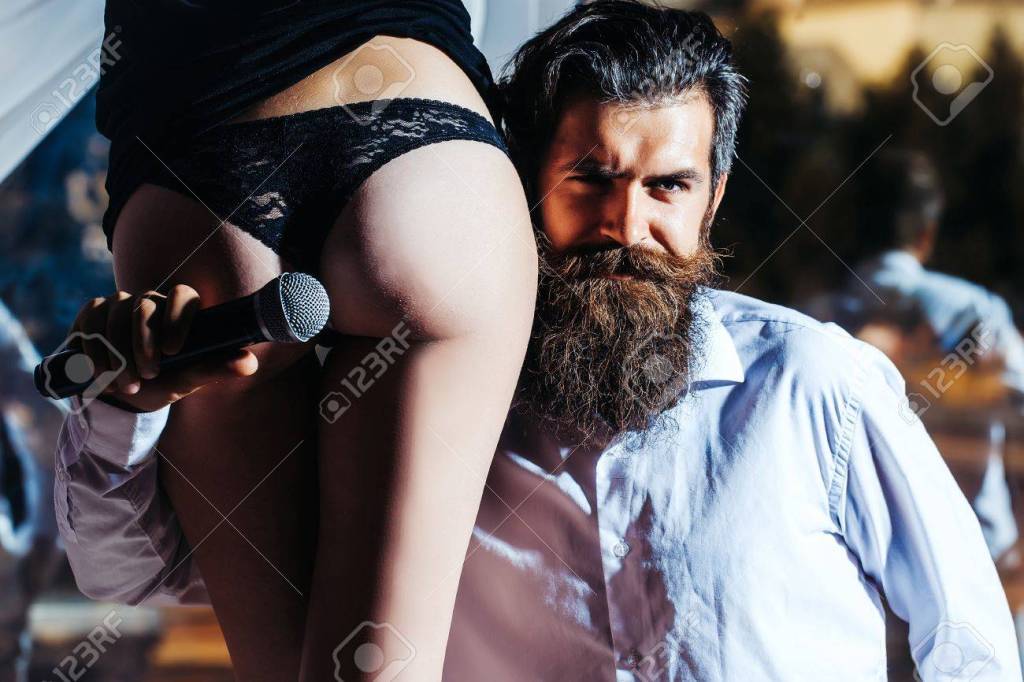 A bearded man holding a microphone to the butt of a woman wearing thong panties.